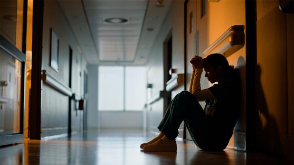 stressed staff member wearing scrubs sitting in hospital corridor with forehead resting on clasped hands