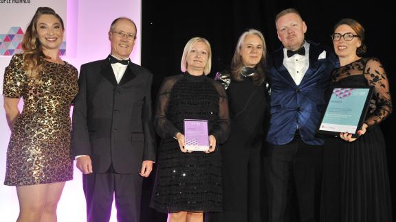 A small group of individuals standing on a stage smartly dressed receiving an award for the HPMA Awards 2023.