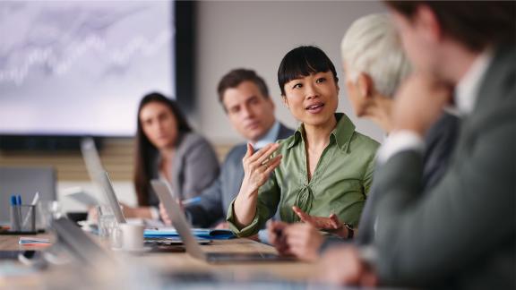 five people seated at a desk in an office with centremost colleague sharing their ideas